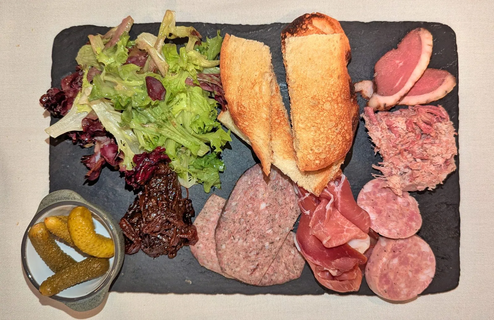 A slate charcuterie board with sliced cured meats, pâté, bread, salad greens, and pickles